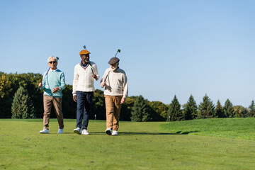 senior interracial friends walking with golf clubs on green field.