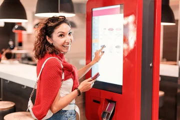 Deurstickers A girl orders food and lunch at a fast food restaurant using a self-service kiosk or a terminal with a screen. Modern commerce equipment © EdNurg