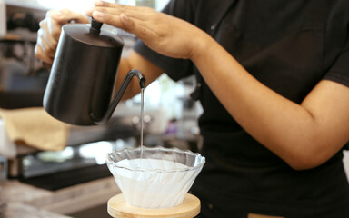 A female cafe operator wearing an apron pours hot water over roasted coffee grounds to prepare coffee for customers in the shop.