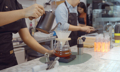 A female cafe operator wearing an apron pours hot water over roasted coffee grounds to prepare coffee for customers in the shop.