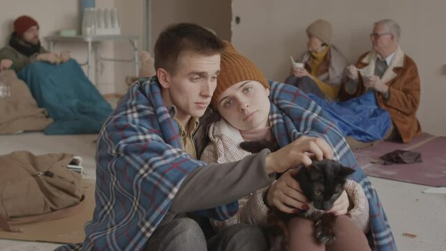 Slowmo portrait of sad young Caucasian couple of refugees petting homeless black cat and looking at camera, staying in cold uncomfortable asylum with other refugees