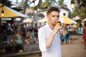 Fototapeta premium Man drinking beer while standing in the summer bar. Male holding alcohol drink at outdoor pub. Young man enjoying drinks at beach bar. Glass of crafting beer. Enjoy life style concept