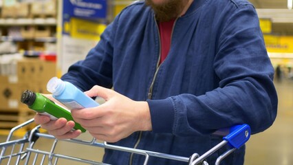 A man in a store chooses a shower gel, carefully reads the composition, then puts one bottle on the shelf and puts the other in a shopping cart for purchase. Choosing a shampoo in a supermarket