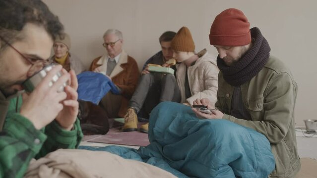 Slowmo portrait of bearded Caucasian refugee man using mobile phone to connect with family then looking sadly at camera sitting on floor of cold asylum with other refugees