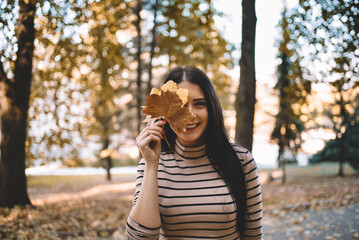 Autumn outdoor portrait of cute smiley women holding autumn leaves near the face.