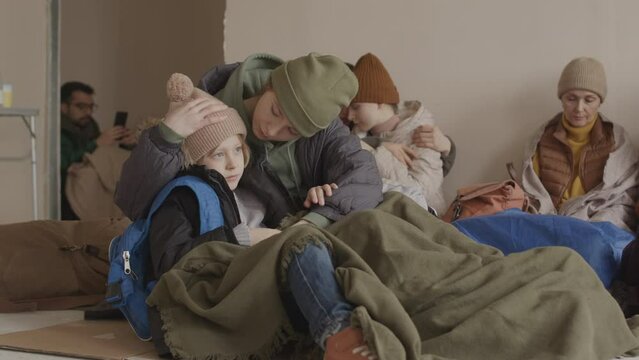 Slowmo of caring young Caucasian woman embracing her 7 year old son sitting close to each other in cold shelter with other refugees, having nowhere to go