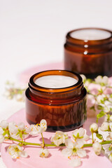Open amber glass jar with cream texture with blooming spring white flowers. Skincare products, natural cosmetic. Beauty concept for face body care