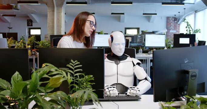 Robot sitting at table in office, typing on keyboard. Woman talking and helping. Android working at computer in coworking space with female. Humanoid as IT worker. Futuristic. Artificial intelligence