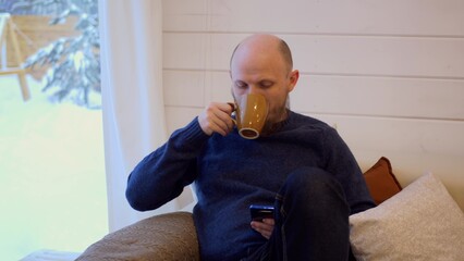 A brutal man with a beard and mustache is sitting at home reading messages on his smartphone and drinking hot coffee. A man's morning starts with coffee and news on the Internet.