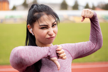 Woman dressed sports suit showing muscles biceps triceps while training running jogging doing sport at the stadium treadmill. Athlete posing body, thumb down dislike gesture sign