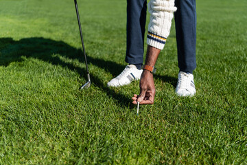 cropped view of african american senior man placing golf tee on green lawn.