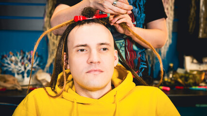 Fototapeta na wymiar Unrecognizable person makes hairstyle for young man in salon. Professional hairstylist makes brown dreadlocks for client.
