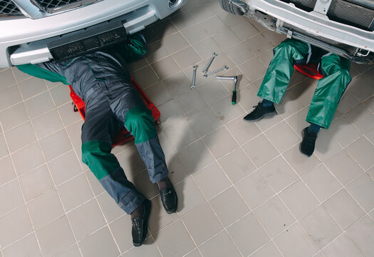 Mechanics in uniform lying down and working under car at the garage.