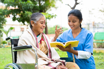 Nurse and senior woman on wheel chair laughing by reading book or novel at park - concept of...