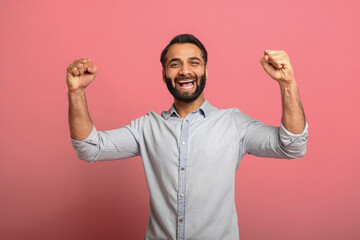 Lucky one. Excited Indian guy celebrating victory and gesturing YES with fists raised on pink background. Overjoyed multiracial bearded man in casual jeans shirt winning