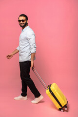 Full length view of the smiling positive Indian man carrying luggage and stepping somewhere while looking at the camera, isolated on pink. Traveling concept