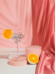 Martini glass with ice and orange fruit, palm leaf shadow and podium on pink background. Pastel...
