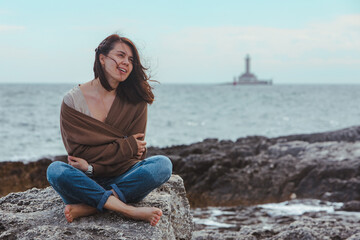 woman sitting by rocky sea beach in wet jeans lighthouse on background
