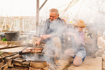 granddaughter helps grandfather cook barbecue. in the smoke from the fire. 