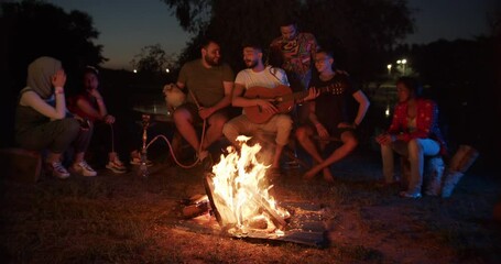 A group of international people friends sitting by the fire playing guitar and singing songs. The dog is sitting on a bench next to a man. Hookah.
