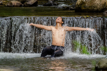 Asian man relaxing with freedom open arms in waterfall in tropical nature. Wellness spa concept in holiday nature.