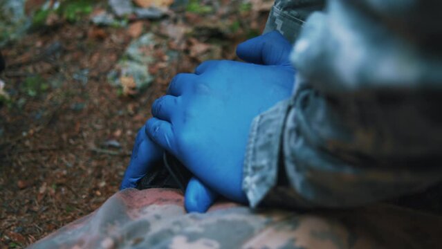 Hands military medic with gloves presses the wound with his hands. Soldiers lying on ground. Blood around