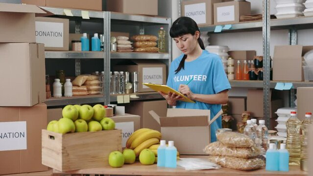 Caucasian Female Volunteer In Blue Shirt Writing On Clipboard While Working At Food Bank Warehouse. Young Woman Doing Inventory Of Goods On Shelves.