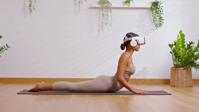 Asian woman using VR goggles practicing yoga cobra pose workout in Metaverse online community.Yoga training class for health with future digital tech virtual reality exercise in Metaverse by her self