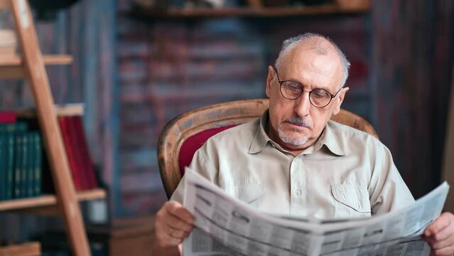 Pensive 70s mature grandfather reading paper publication newspaper text journal on vintage armchair 