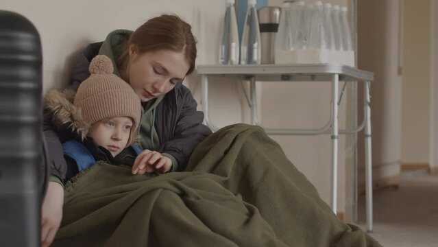 Slowmo of upset young Caucasian refugee woman and her little son sitting close to each other in shelter wearing old warm clothes, having nowhere to go