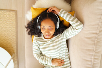 Calm African-American female listening to music while lying down on couch in living room, overhead