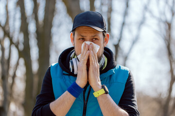 A man with a pollen allergy stands outside in a park. An athlete stops running practice due to a runny nose, blows his nose in a handkerchief. Illness interferes with exercise. - obrazy, fototapety, plakaty