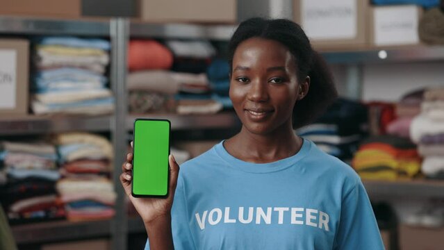Portrait view of the multiracial woman holding smartphone with green mock up screen and showing it to the camera while standing at the donation center. Volunteers concept