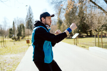 Outdoor workout. Handsome young man does warm-up exercises before running while standing still, preparation for workout, boxing with hands, motivational music in headphones.