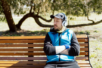 A man wearing a helmet, fleece and a vest is sitting on a park bench listening to music on wireless...