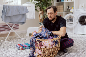 Portrait of man sitting in laundry room on floor by wicker basket of clothes with a phone in hand. The boy is sorting shirts checking the internet for pictograms, washing machine instructions.