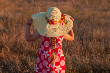 Adorable littAdorable little girl in straw hat pink summer dress with spikelets in hand in whle girl straw hat pink summer dress with spikelets in hand in wheat field back view. Long blonde hair child