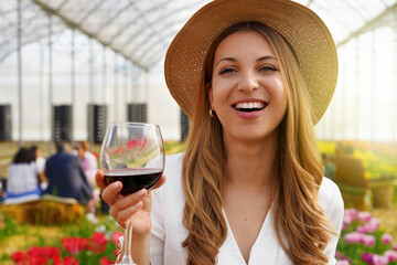 Close-up of young woman looking at camera and holding glass of wine with people among flowers field...