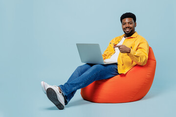 Full body happy young man of African American ethnicity wear yellow shirt sit wearing bag chair hold use work point finger on laptop pc computer isolated on plain pastel light blue background studio