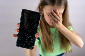 Scared little girl with emotional expression showing broken smartphone. Portrait frustrated kid...