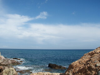 Mar con poco oleaje y espumacon rocas de playa en primer plano  en los laterales y nubes en el cielo