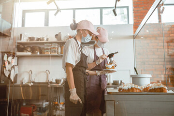 Woman in mask holds cake with burnt cream near colleague with gas burner in bakery