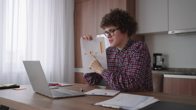 A curly man with glasses at his desk shows a picture with graphs in a laptop camera