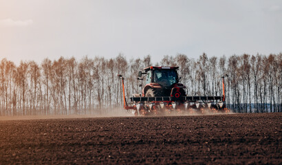 Fototapeta premium Spring sowing season. Farmer with a tractor sows corn seeds on his field. Planting corn with trailed planter. Farming seeding. The concept of agriculture and agricultural machinery.