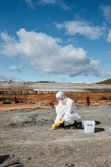 Researcher in hazmat suit and gloves sitting on abandoned area and taking sample while studying toxic soil