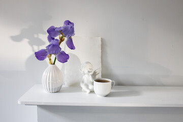 Blue iris in a corrugated vase with a piece of craft paper on a white table. Figurine of the angel. Cup of tea. Place for text