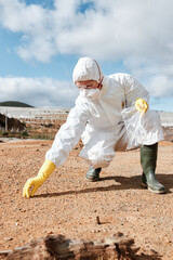 Environmental researcher in mask and safety goggles taking sample of soil and putting it into lastic bag on polluted land