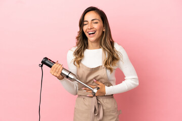 Young woman using hand blender over isolated pink background smiling a lot