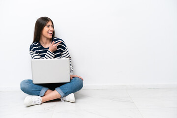Young caucasian woman with a laptop sitting on the floor isolated on white background pointing finger to the side and presenting a product