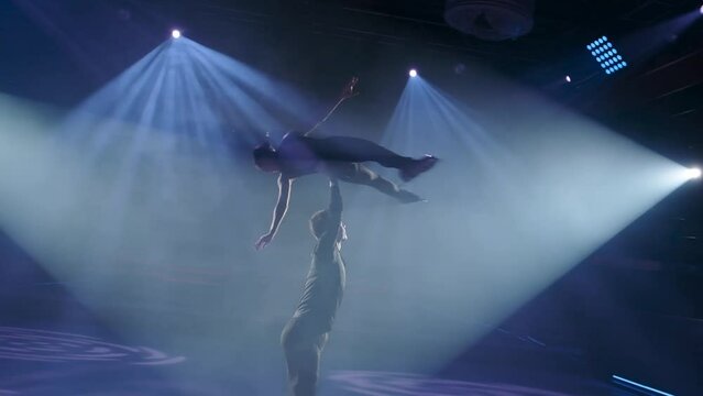 Figure skaters pair perform stationary rotational overhead lift. Man rotates woman high up in the air on one straight hand,her legs and arms outstretched.Risky forbidden support,slow motion,close-up.